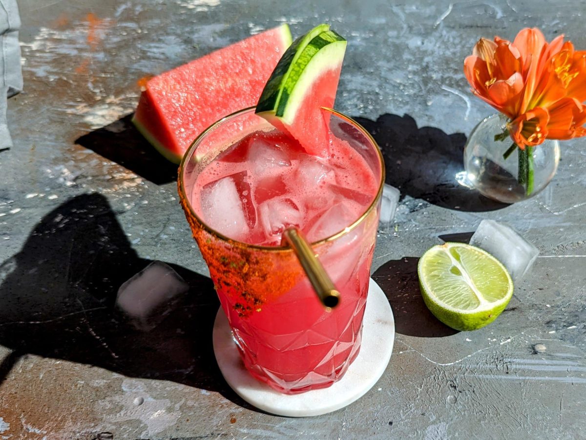 Ein glas mit dem pinken Watermelon-Paloma auf einem grauen Tisch mit rötliche rkleiner Pflanze, Eiswürfeln, halber Limette und grauer Serviette.