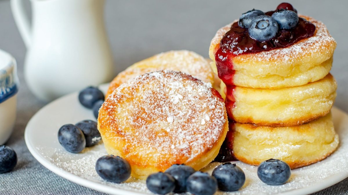 Einige Quarkpfannkuchen mit Blaubbersauce und frischen Blaubeeren auf einem weißen Teller und grauem Untergrund.