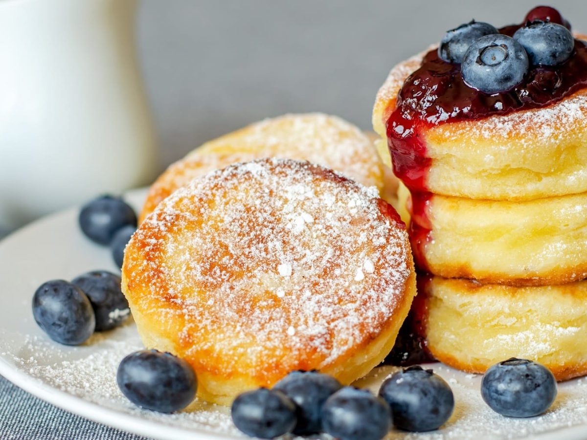 Einige Quarkpfannkuchen mit Blaubbersauce und frischen Blaubeeren auf einem weißen Teller und grauem Untergrund.