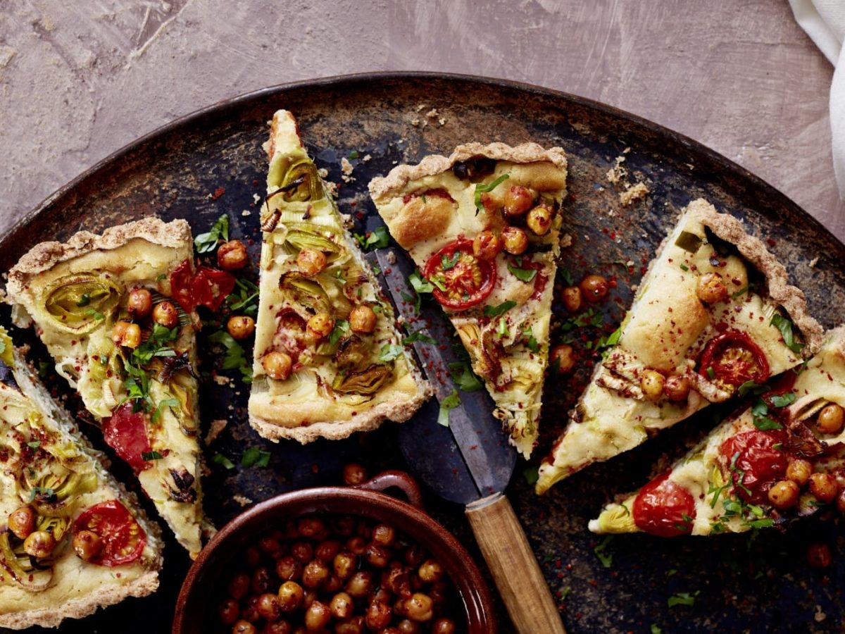 Geschnittene Quiche mit Cashew-Guss und Röstkichererbsen auf einem dunklen Teller, wovon nur die Hälfte zu sehen ist. Untergrund braun.