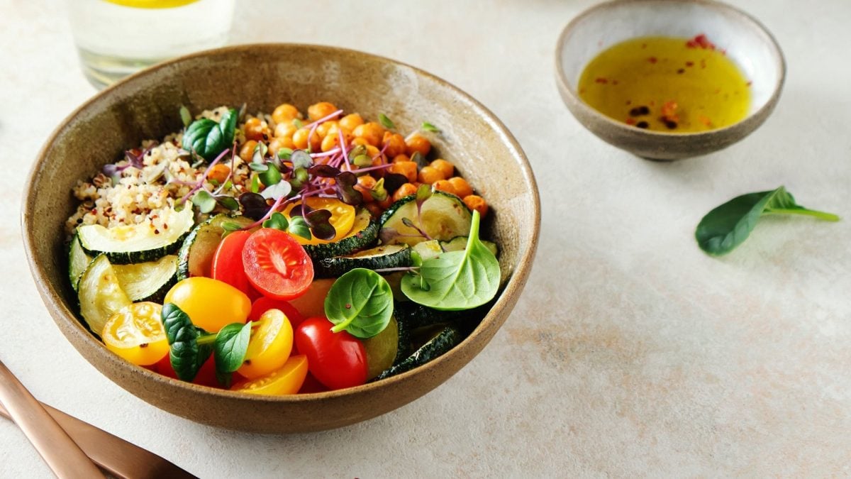 Eine Quinoa-Kichererbsen-Bowl mit Tomaten, Zucchini, Babyspinat und Microgreens, daneben eine Schale mit hellem Dressing.