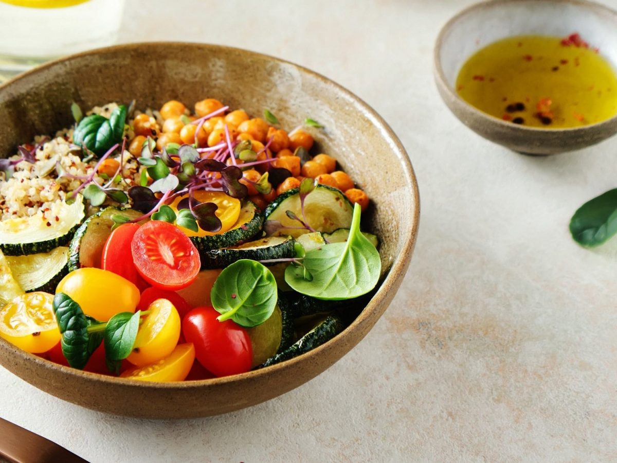 Eine Quinoa-Kichererbsen-Bowl mit Tomaten, Zucchini, Babyspinat und Microgreens, daneben eine Schale mit hellem Dressing.