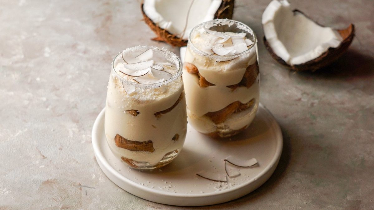 Zwei Gläser Raffaello-Tiramisu mit Kokosflocken und Kokosraspeln auf einem runden Teller, im Hintergrund eine aufgebrochene Kokosnuss.