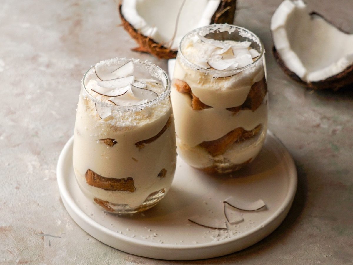 Zwei Gläser Raffaello-Tiramisu mit Kokosflocken und Kokosraspeln auf einem runden Teller, im Hintergrund eine aufgebrochene Kokosnuss.