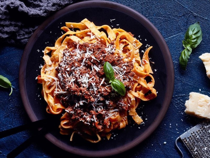 Ragù alla bolognese mit Tagliatelle, Parmesan und Basilikum auf einem Teller in der Draufsicht, daneben liegen Parmesanstücke, eine Käsereibe und Basilikumblätter.