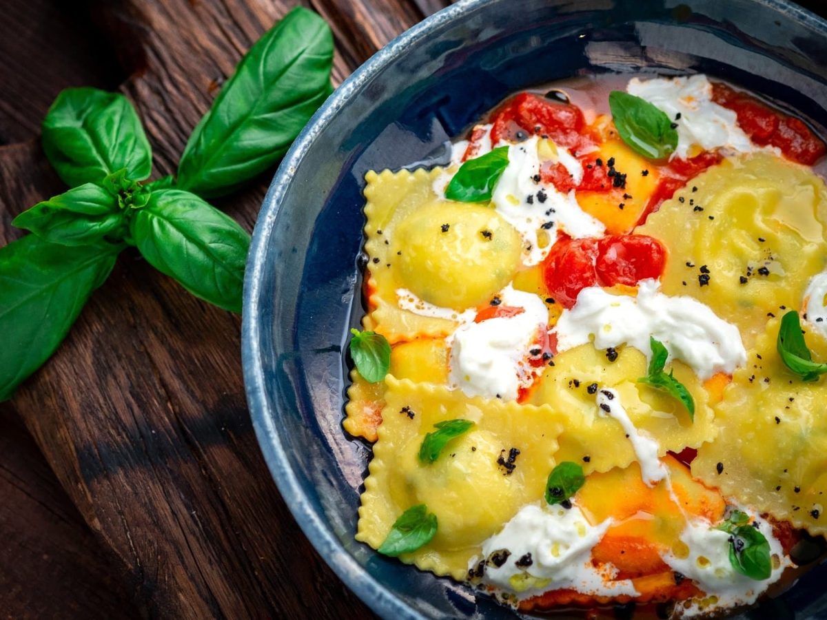 Draufsicht: Eine dunkle Pfanne Ravioli alla Caprese liegt auf einem dunklen Untergrund. In der Pfanne sind Nudeln, Tomaten, Basilikum und weiße Käse. Drum herum liegen frische Blätter Basilikum.