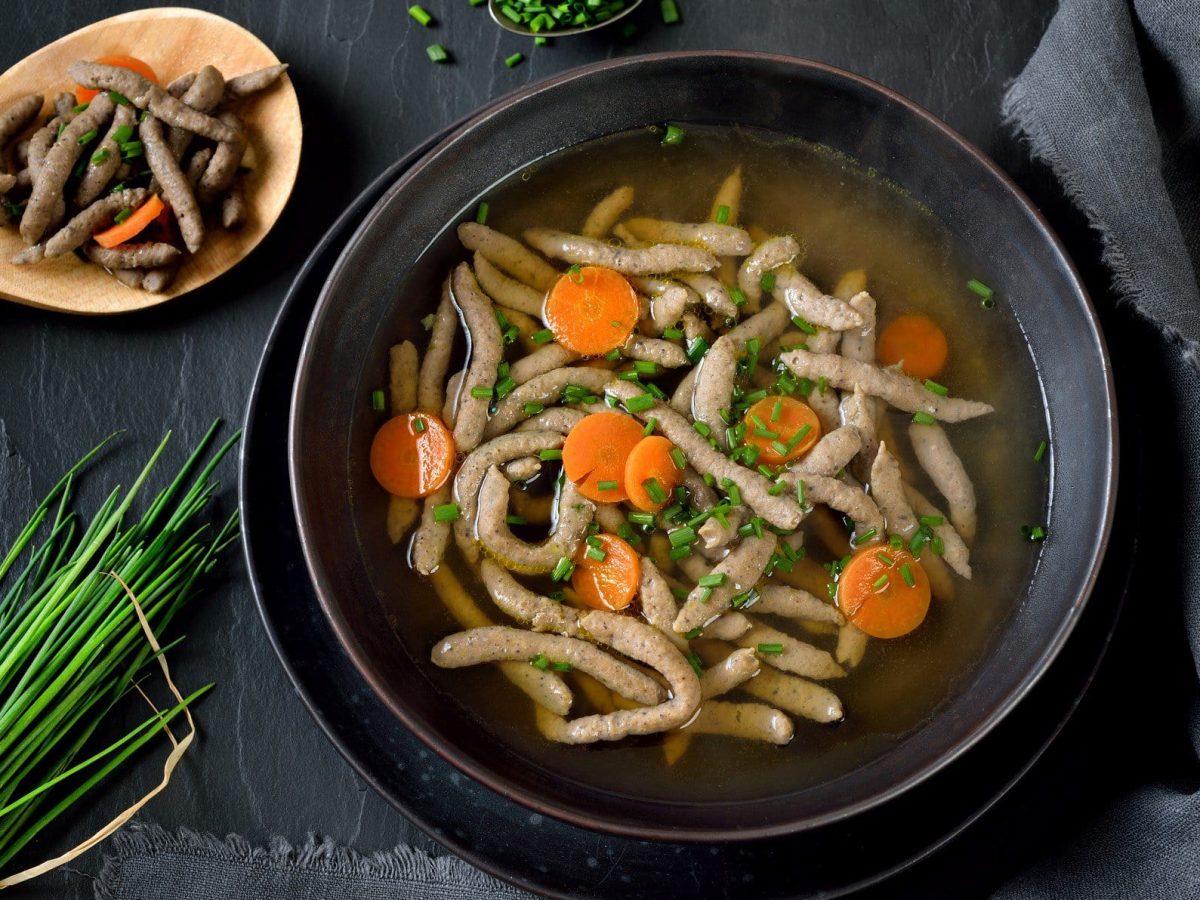 Draufsicht: Auf einem schwarzem Untergrund steht ein dunkler Teller Rindersuppe mit Leberspätzle. Drum herum liegen frischer Schnittlauch un ein Kochlöffel, auf dem Leberspätzle mit Karottenscheiben liegen.