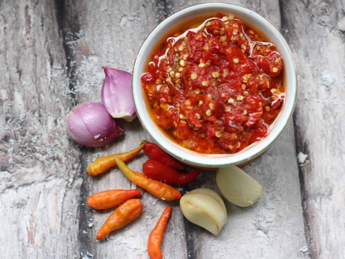 Eine weiße Schale Sambal Bawang indonesisches Sambal auf hellem Holz. Im Bild Schalotten, rote Chilischoten und geschälte Knoblauchzehen. Draufsicht.