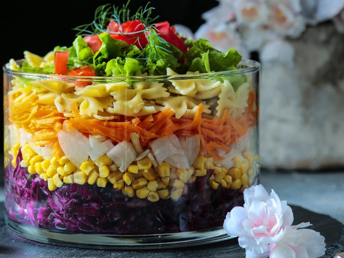 Eine große Glasschüssel mit dem geschichteten Salat auf einer schwarzen Schieferplatte mit weißen Blüten und Knospen im Hintergrund.