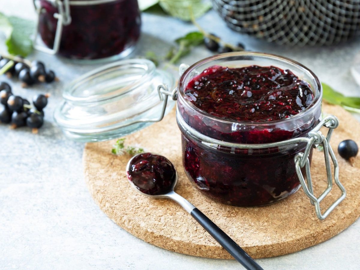 Draufsicht, schräg: Ein Einmachglas Schwarze Johannisbeeren-Marmelade steht auf einem Brettchen auf einem hellen Untergrund. Drum herum liegen schwarze Johannisbeeren und Blätter.