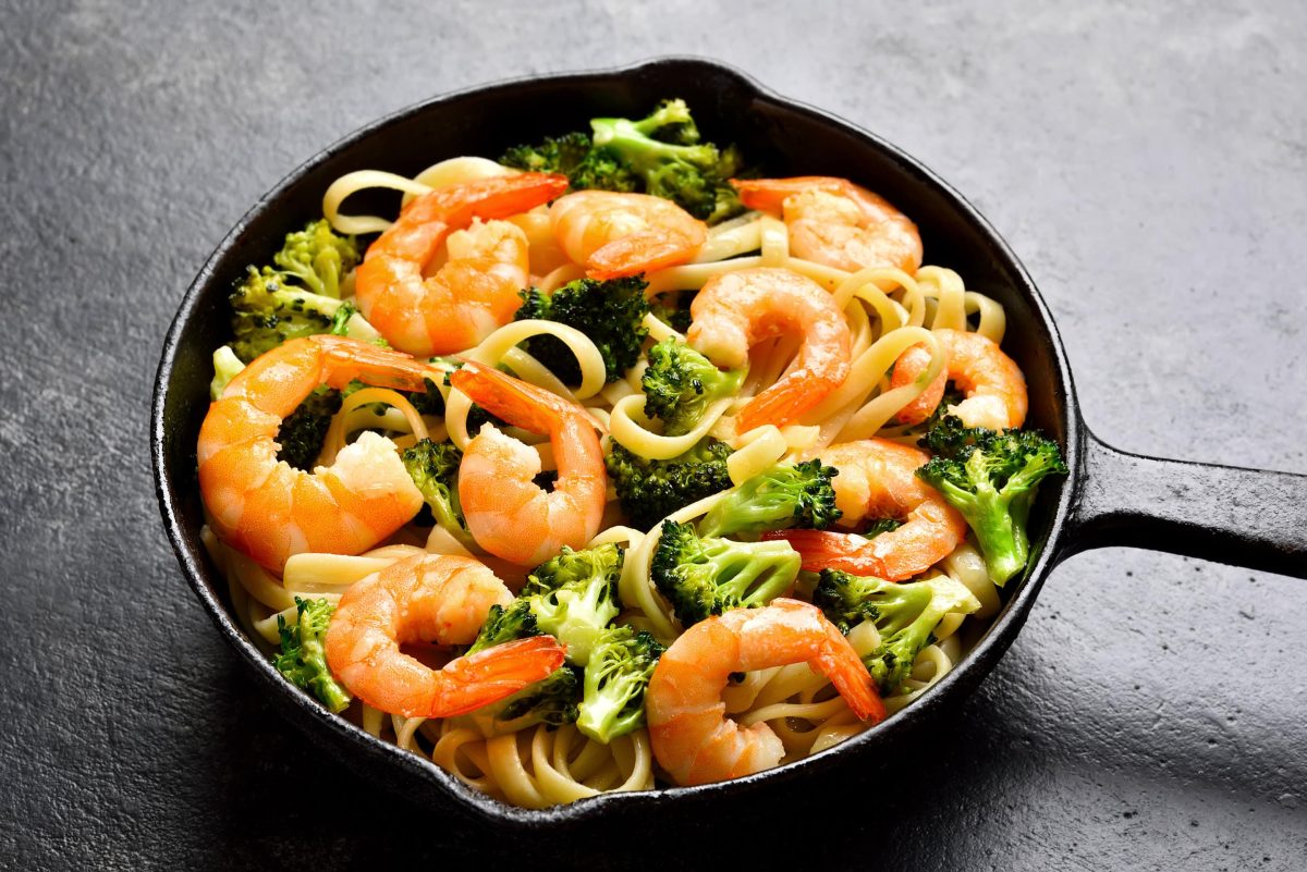 Draufsicht: eine schwarze Pfanne mit Shrimps-Brokkoli-Pasta liegt auf einem grauem Untergrund.