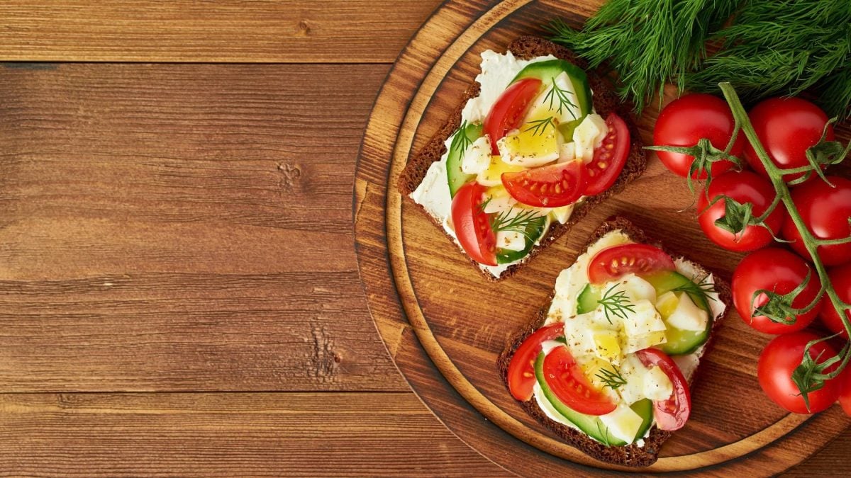 Draufsicht: Auf eine runden Holzbrettchen liegen zwei Sommer-Sandwiches. Die sind mit Frischkäse, Tomaten und Gurken belegt. Daneben liegen frischen Rispentomaten.