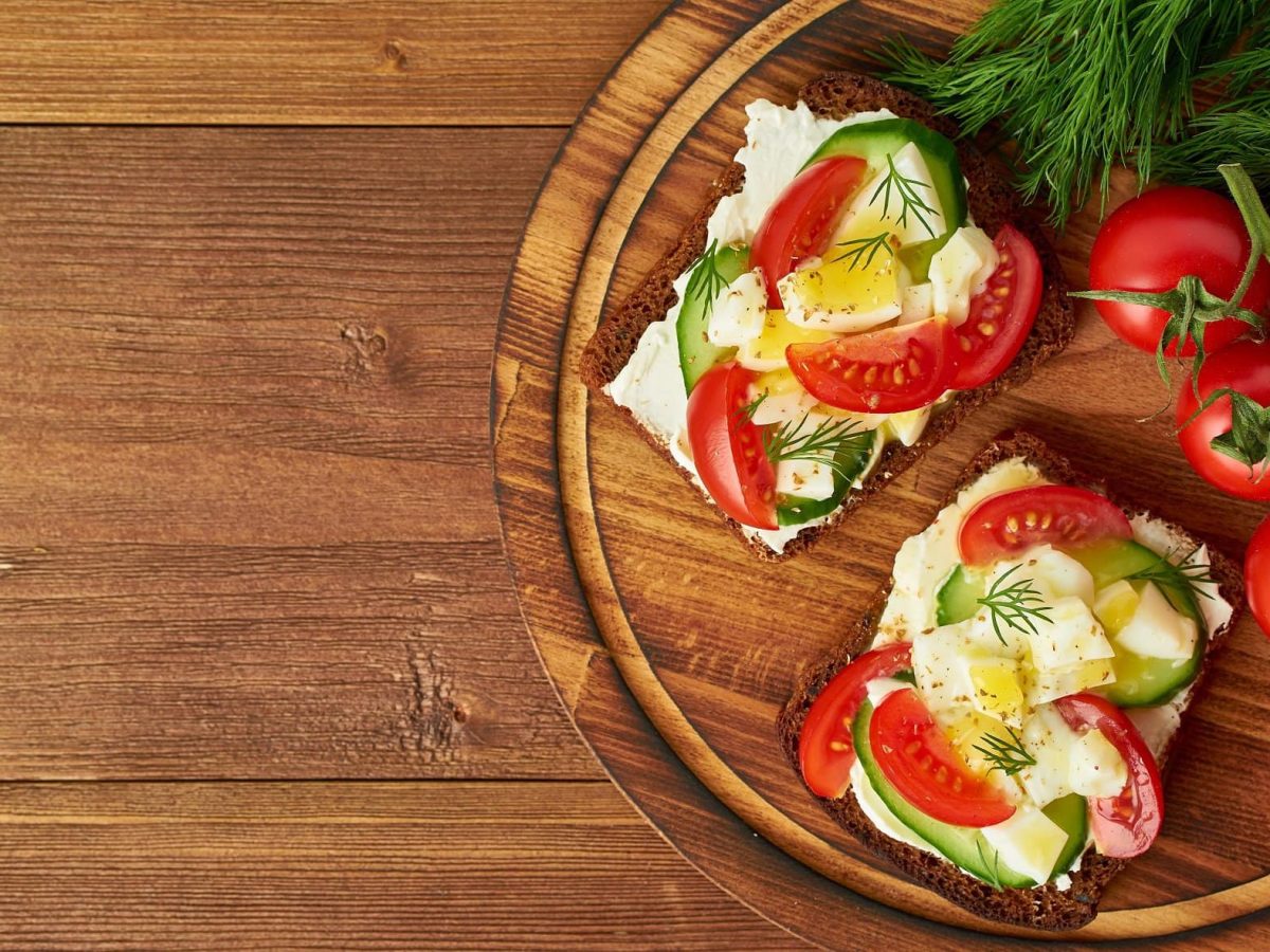 Draufsicht: Auf eine runden Holzbrettchen liegen zwei Sommer-Sandwiches. Die sind mit Frischkäse, Tomaten und Gurken belegt. Daneben liegen frischen Rispentomaten.