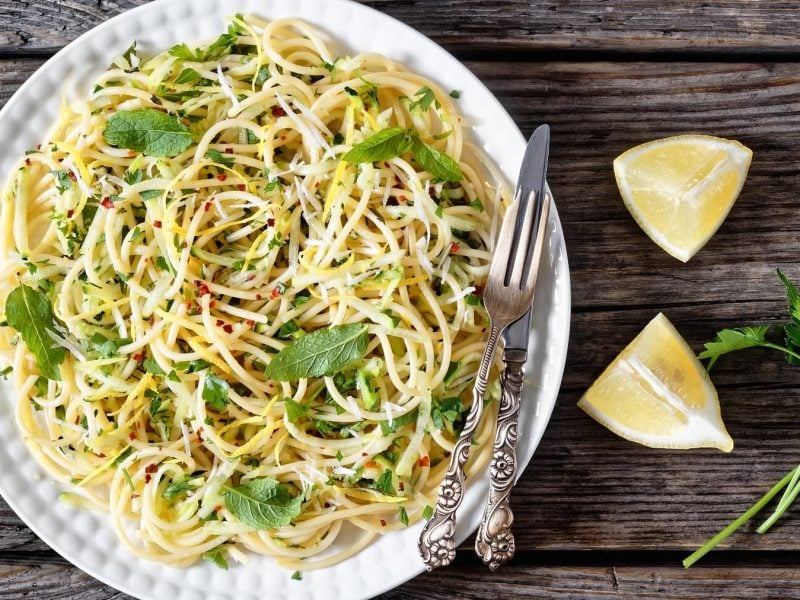 Ein weißer Teller mit der Pasta auf einem dunklen, rustikalem Holztisch. Neben der Spaghetti Carrettiera liegen Zitronenecken und Petersilie.