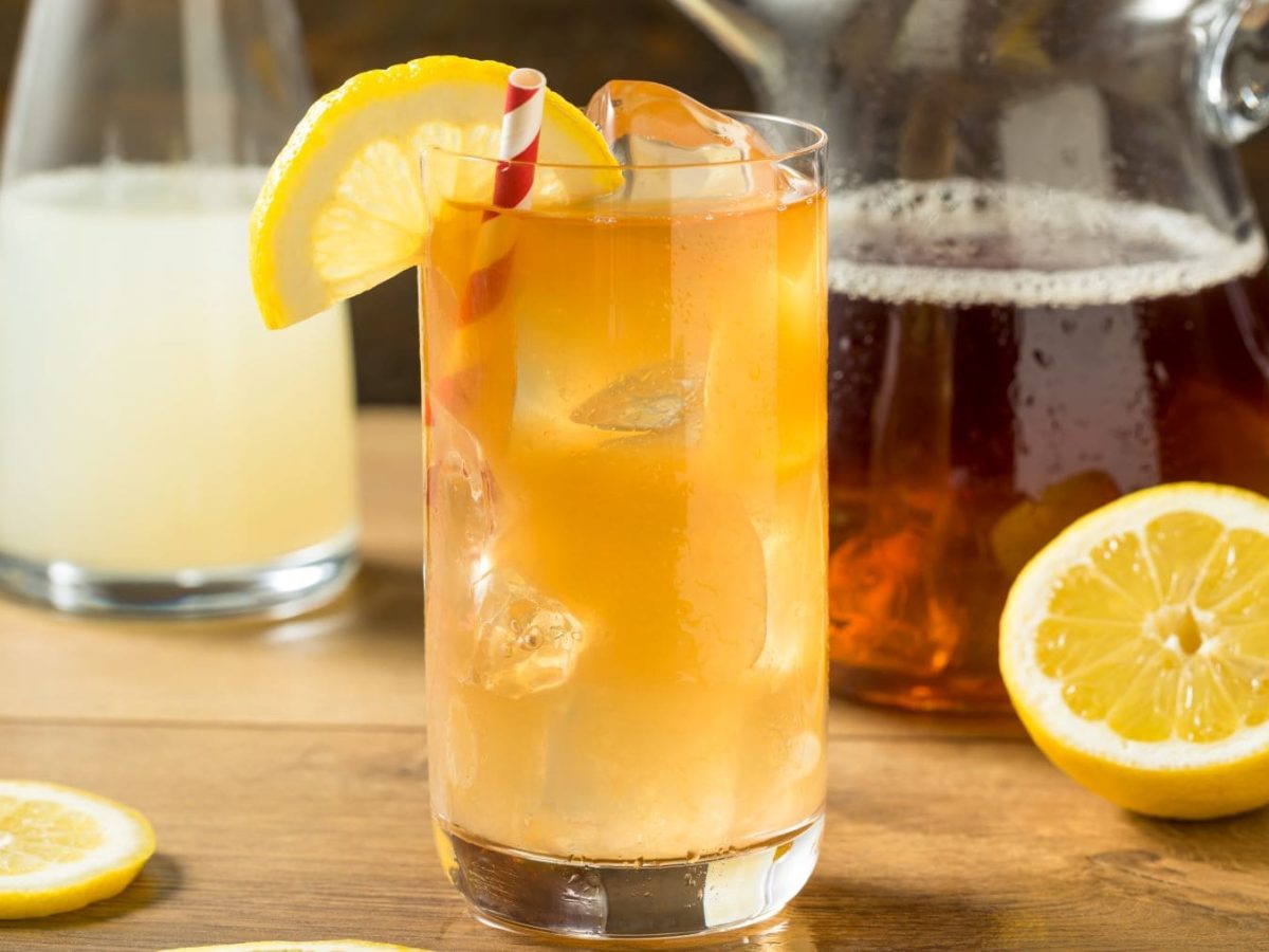 Vodka Spiked Arnold Palmer alias Jon Daly Cocktail mit Zitronenscheibe im Longdrink-Glas auf Holz. Im Bild Zitronen, Zitronenscheibe und je eine Karaffe mit Limonade und Eistee. Frontalansicht.