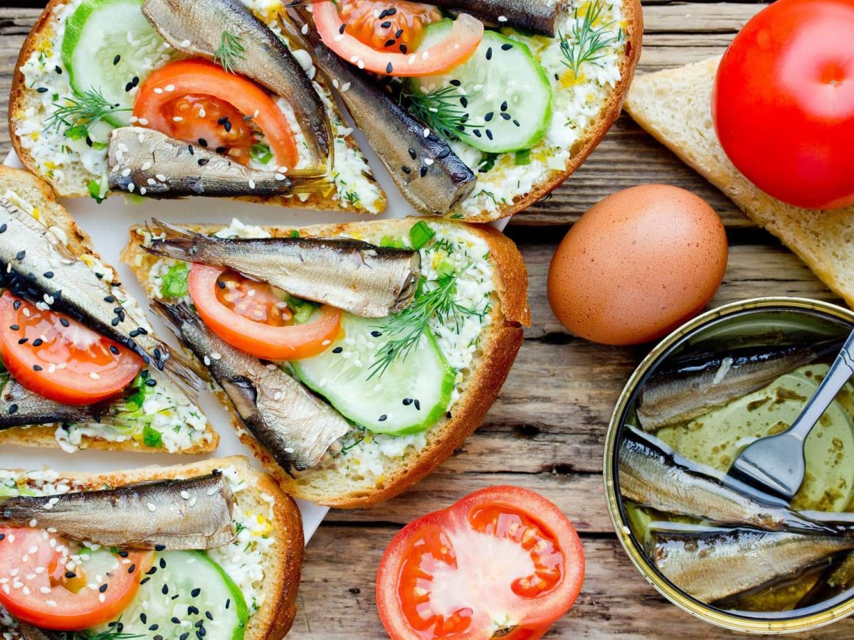 Draufsicht: Sprotten-Sandwich mit Tomaten, Gurken und schwarzem Sesam liegen auf einem Untergrund aus Holz.