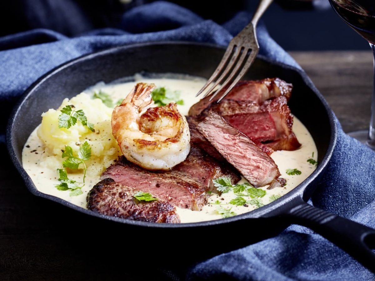 Ein Steak mit Sauce und Garnelen n einer schwarzen Pfanne auf einem dunkelblauen Geschirrtuch. Rechts davon ein Glas Rotwein auf einem hölzernen Hintergrund.