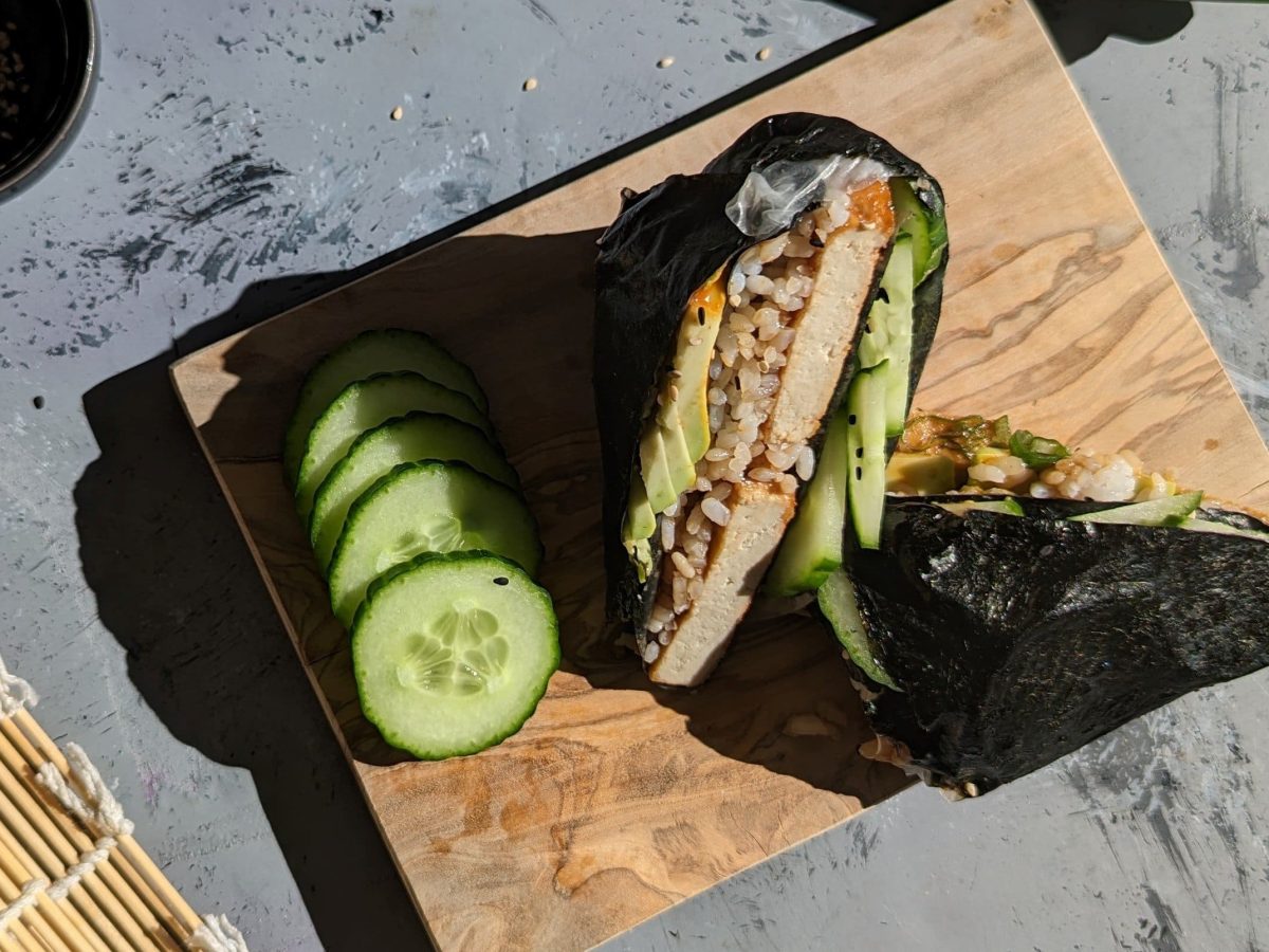 Zwei Sushi-Wraps mit einigen Gurkenscheiben auf einem kleinen Holzbrett und grauem Untergrund. Links daneben eine Sushi-Matte und ein kleines schwarzes Schälchen mit Sesam.