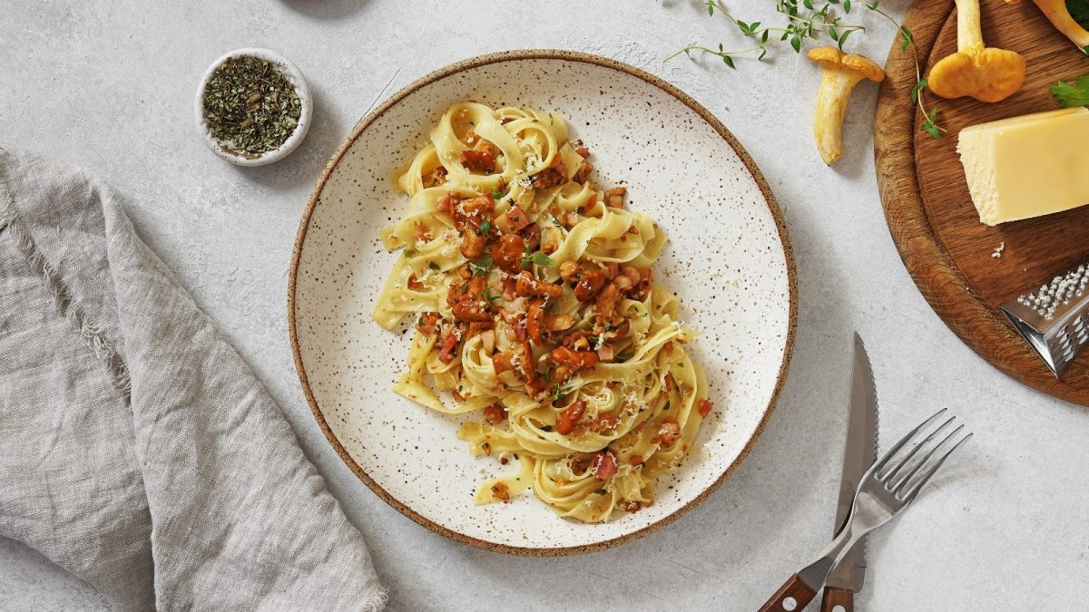 Ein Teller mit Tagliatelle mit Pfifferlingen und Pancetta auf einem weißen Tisch.