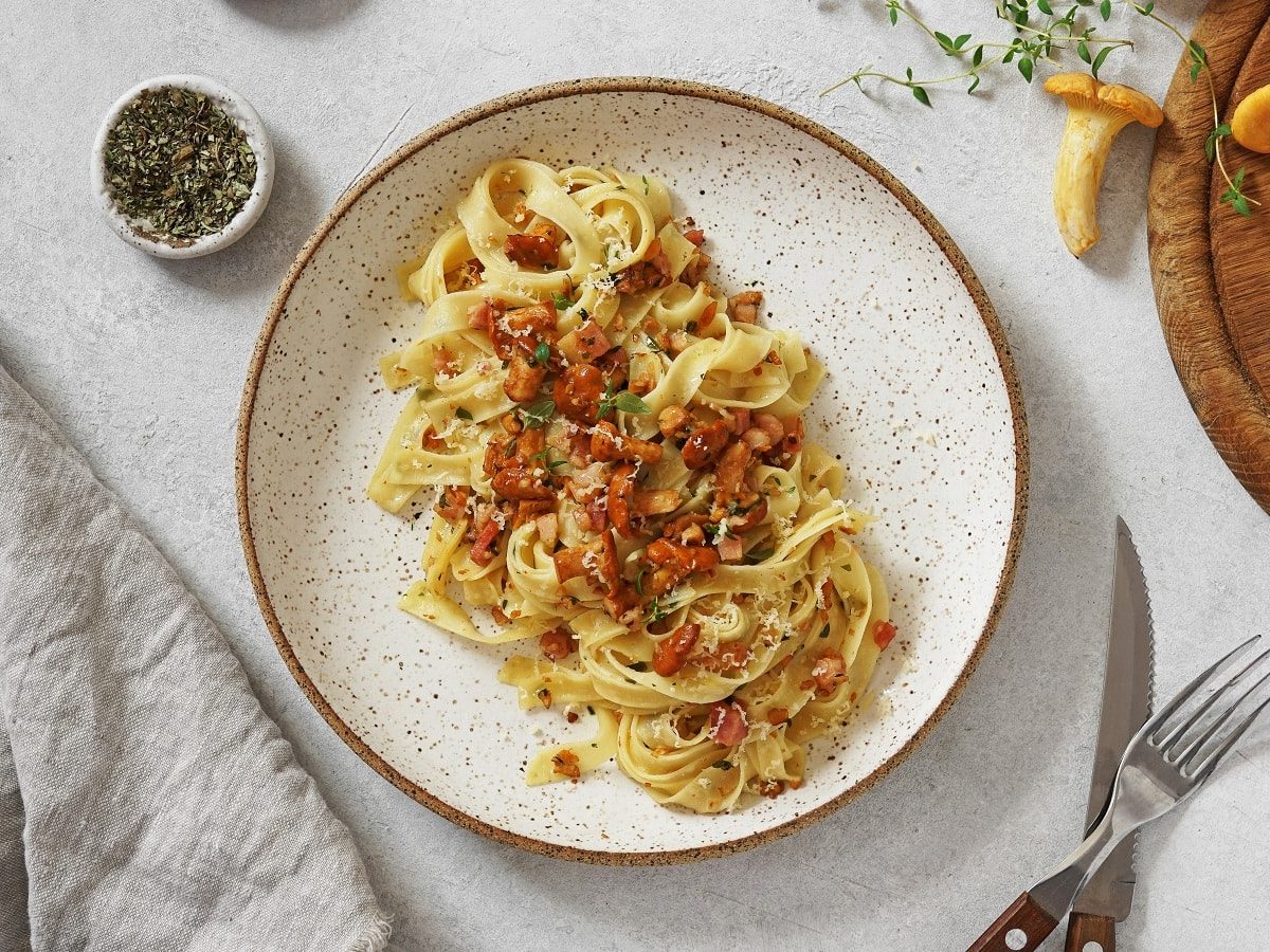 Ein Teller mit Tagliatelle mit Pfifferlingen und Pancetta auf einem weißen Tisch.