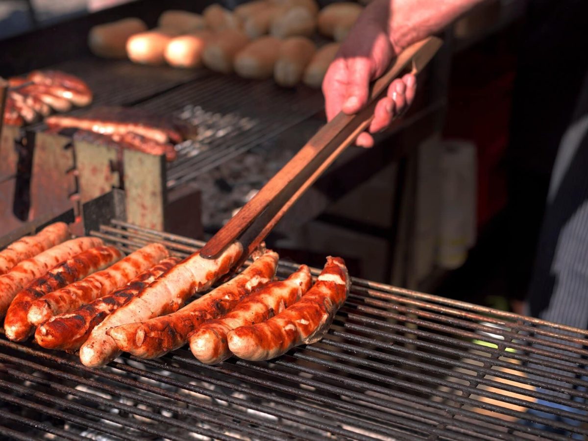 Frontalsicht: Ein Holzkohlegrill mit Thüringer Rostbratwürsten. Eine Hand mit einer hölzernenen Grillzange.