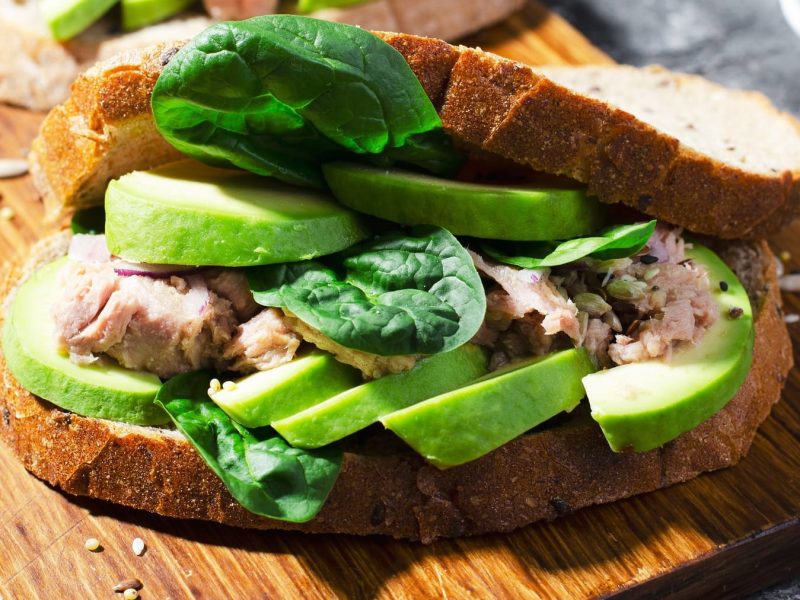 Ein Sandwich mit Avocado, Thunfisch und Babyspinat auf einem Holzbrett und grauem Untergrund.