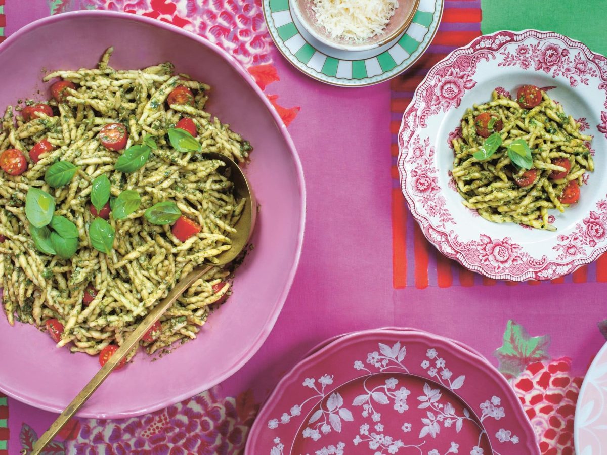 Nudeln mit Tomaten und grünem Pesto auf zwei bunt gemusterten Tellern, die auf pinkem Untergrund stehen, Draufsicht. Bild aus dem Kochbuch "Toskana".