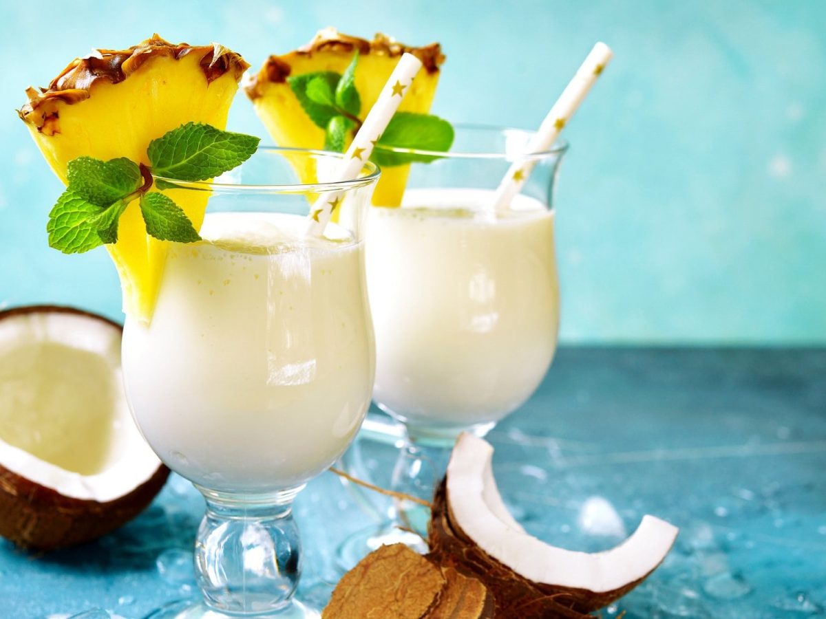 Zwei Gläser vegane Piña Colada, dekoriert mit einer Ananasscheibe und Minze, darin ein Strohhalm, drumherum aufgeschnittene Kokosnüsse.