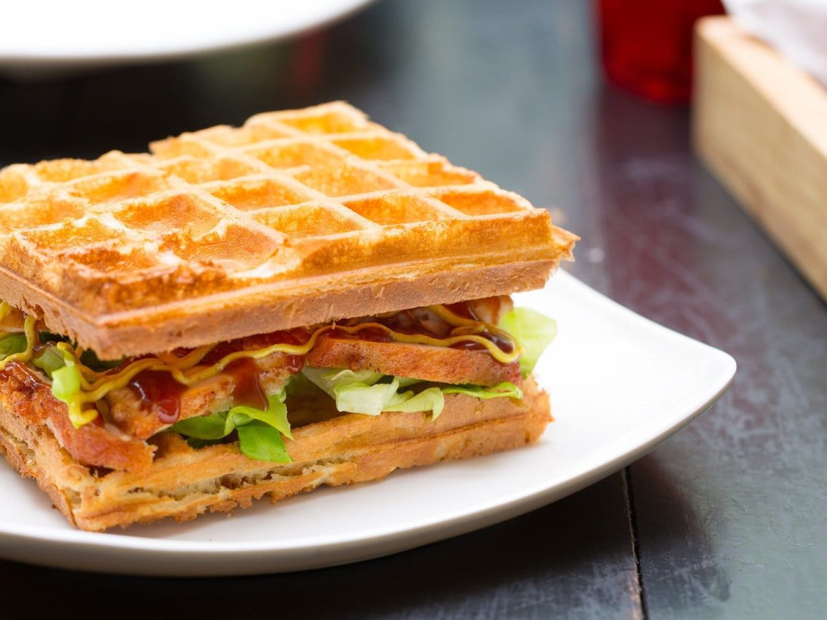 Ein Waffel-Sandwich belegt mit Räuchertofu, Salat, Tomate und Gurke, darauf eine gelbe und rote Sauce, auf einem Teller.