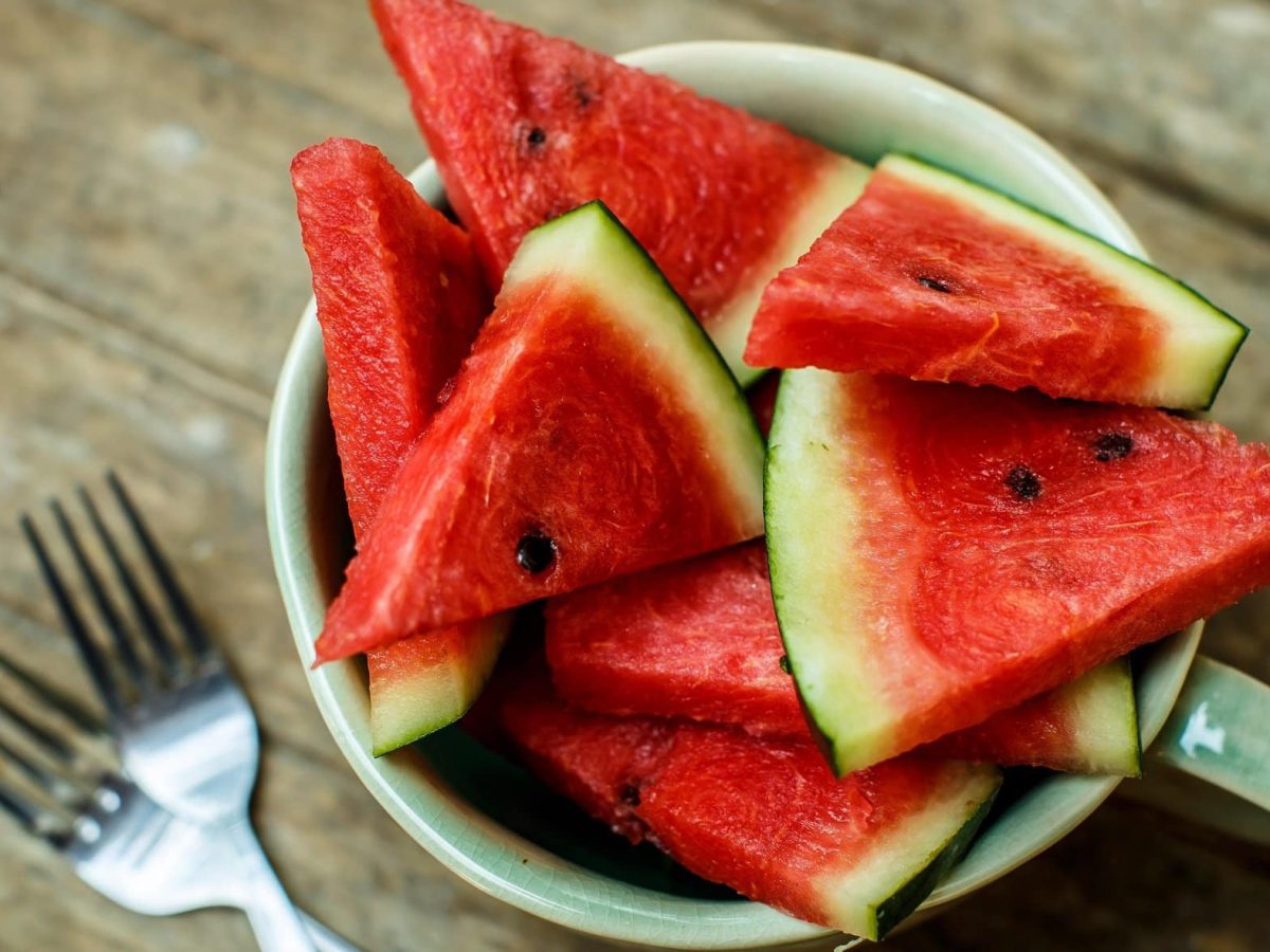 Frontalsicht: Auf einem Untergrund aus Holzplanken steht eine weiße Schüssel mit angeschnittener Wassermelone.