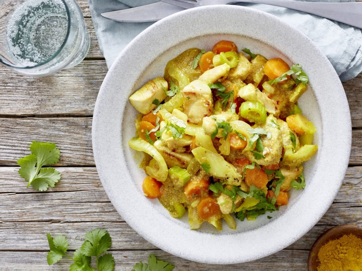 Draufsicht: Amaranth-Fenchel-Pfanne mit Huhn in weißem Teller auf Holzuntergrund, daneben ein Glas und Kräuter verteilt.