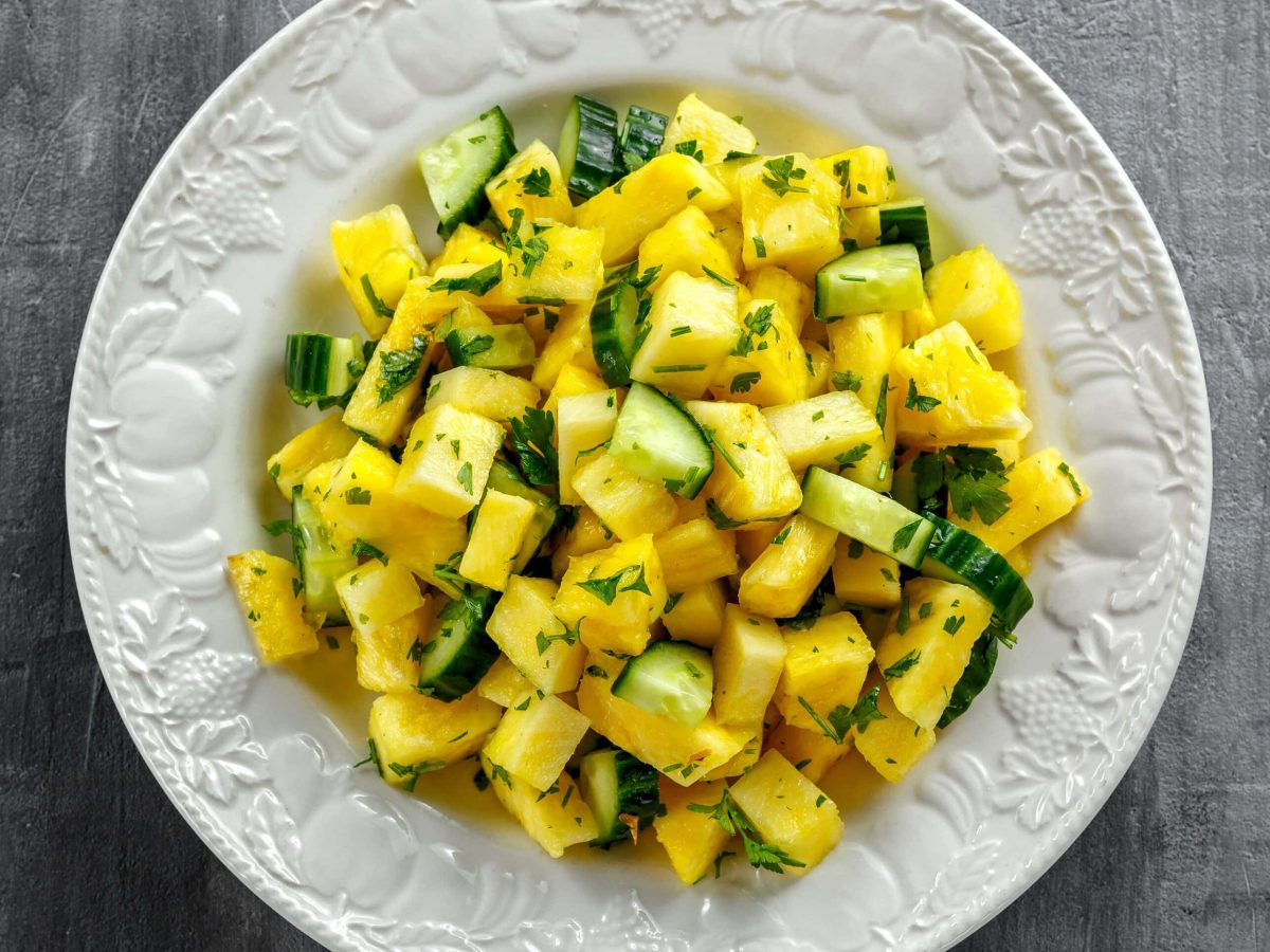 Ein weißer Teller mit dem Ananas-Gurken-Salat auf grauem Untergrund direkt von oben fotografiert.