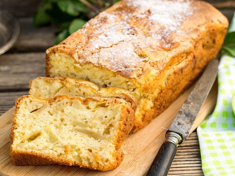 Ein saftiger Apfel-Kastenkuchen auf einem hellen Holzbrett und einem dunklen Tisch mit frischen grünen Äpfeln und einer grün-weiß-karierten Tischdecke.