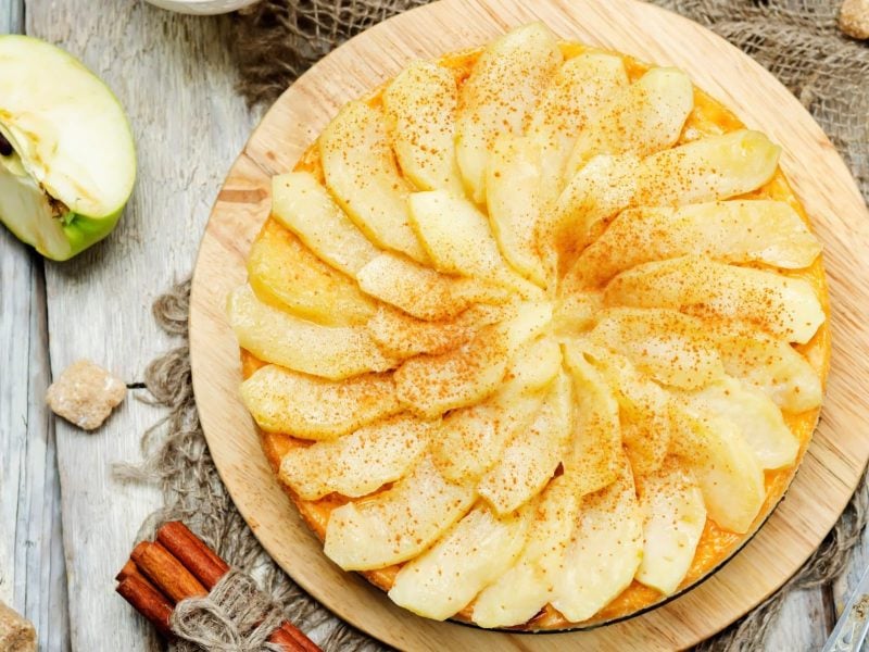 Von-Oben-Ansicht auf gedeckten Apfelmus-Schmandkuchen auf hellem, rustikalem Holz. Daneben liegen Zucker, Apfel und Zimtstangen.