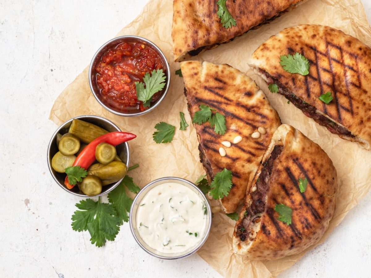 Vegane Arayes auf Backpapier auf einem weißen Hintergrund. Links von den Pita-Broten drei Schälchen gefüllt mit Tahini-Sauce, Salsa und Salzgurken. Alles ist garniert mit frischem Koriander.