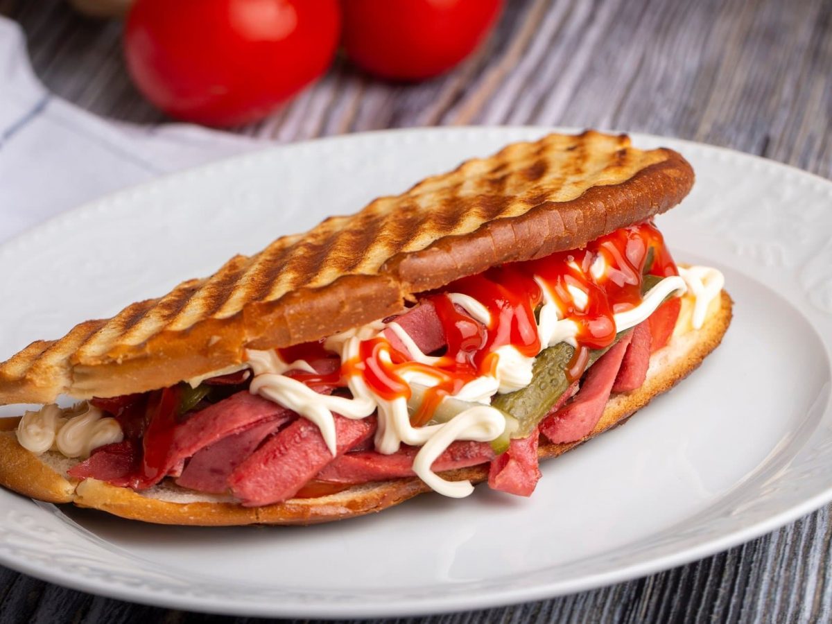 Draufsicht schräg: ein türkisches Sandwich, das Ayvalık Toast mit Sucuk heißt, liegt auf einem weißen Teller. Der Untergrund ist aus Holt und darauf liegen noch zwei Tomaten und ein Tuch.