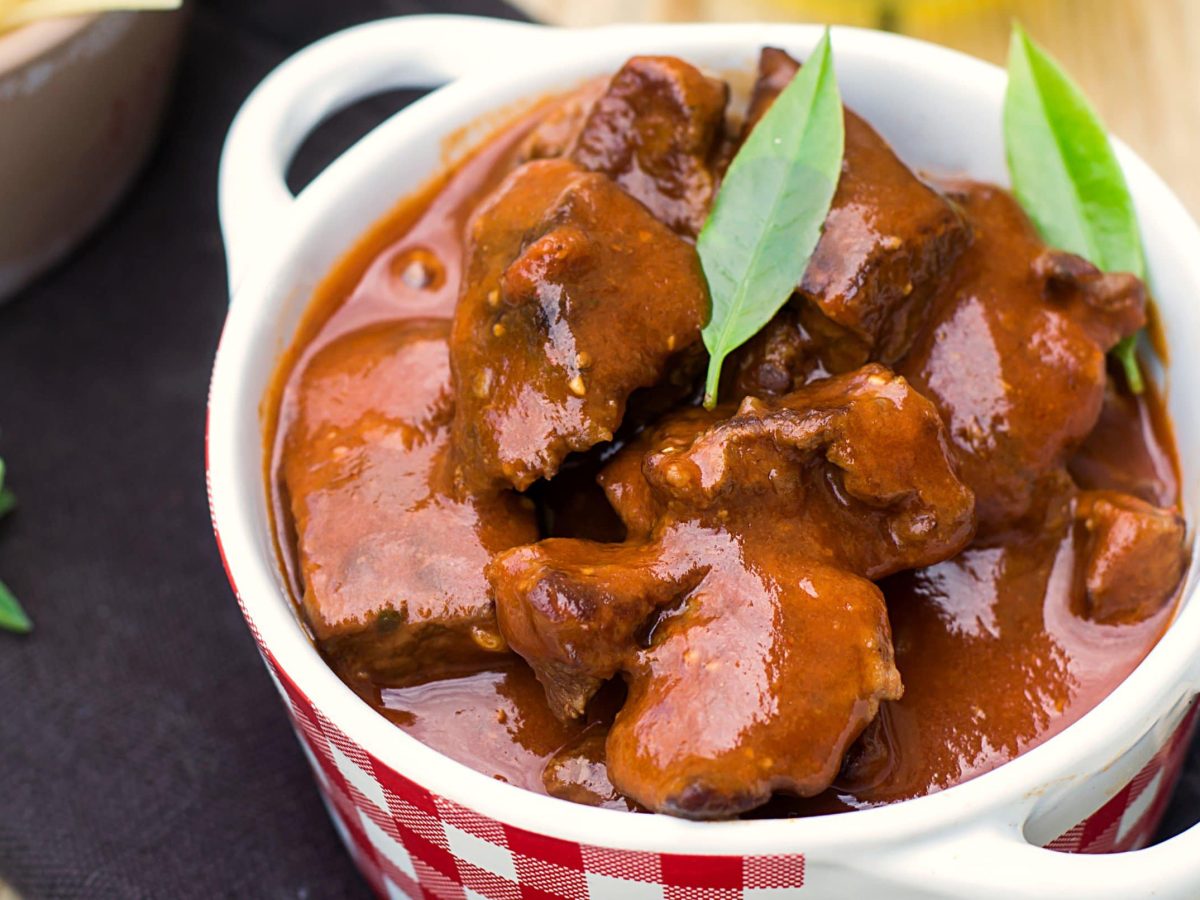 Eine kleine rot-weiß-karierte Suppenschale mit dem Barbecue-Gulasch auf einer schwarzen Tischdecke und einem hellen Holztisch.