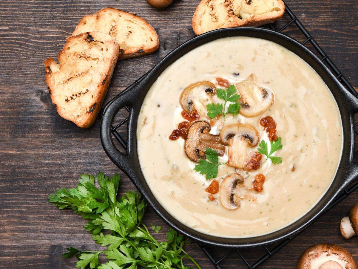 Draufsicht: Dunkler Untergrund, eine dunkle Schüssel bayerische Kümmelrahmsuppe mit Champignons. Daneben liegen frische Petersilie, Brotscheiben und Pilze.