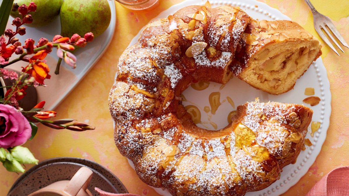 Ein Birnen-Bienenstich-Gugelhupf auf einem weißen Servierteller auf einem orangen Hintergrund. Links davon Blumen und Birnen auf einem weißen Teller. Links unten eine Tasse und rechts oben eine Dessertgabel.