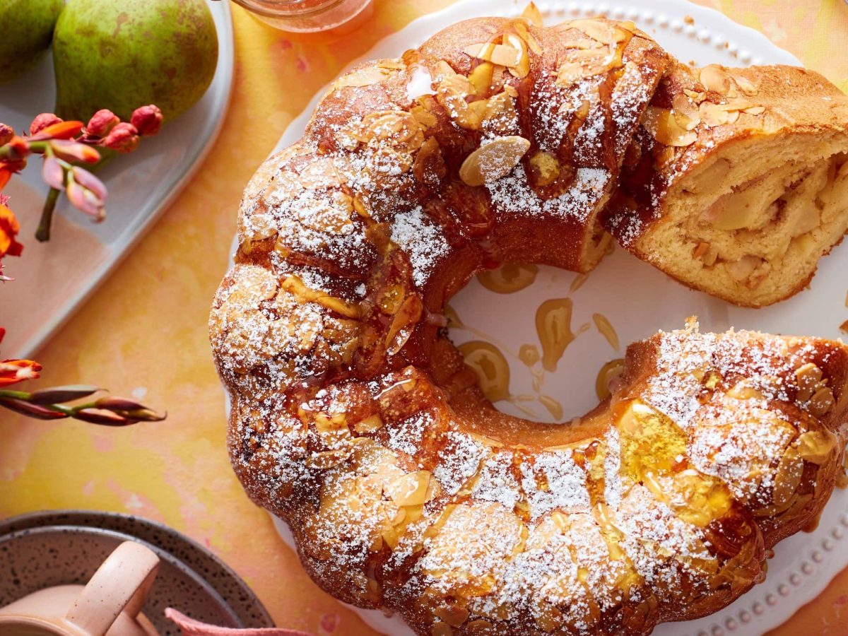 Ein Birnen-Bienenstich-Gugelhupf auf einem weißen Servierteller auf einem orangen Hintergrund. Links davon Blumen und Birnen auf einem weißen Teller. Links unten eine Tasse und rechts oben eine Dessertgabel.