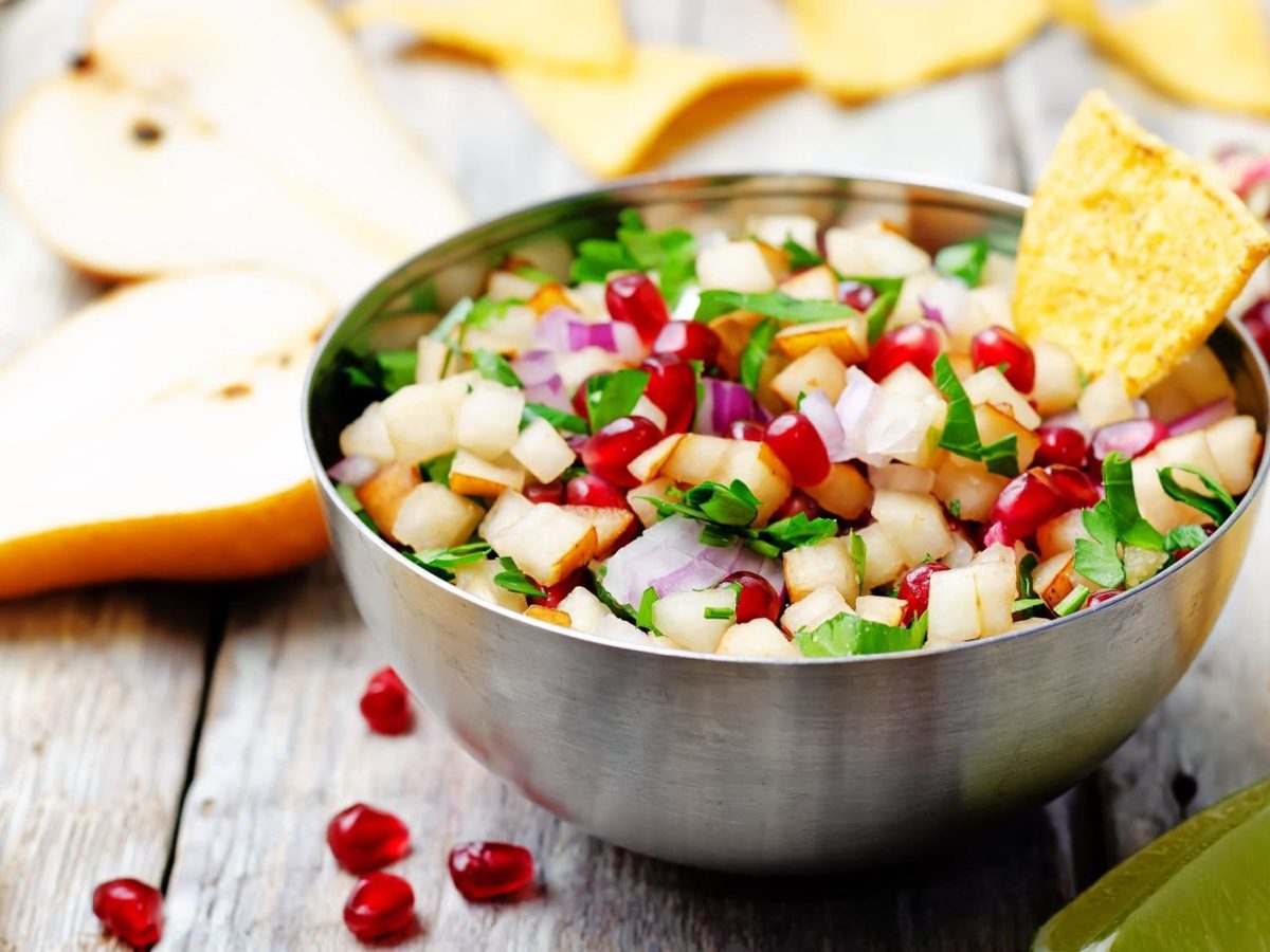 Eine Metallschüssel mit Birnen-Granatapfel-Salsa mit Koriander und roten Zwiebeln, darin steckt ein Nacho-Chip. Drumherum liegen Zutaten wie halbierte Birnen und ein aufgeschnittener Granatapfel.