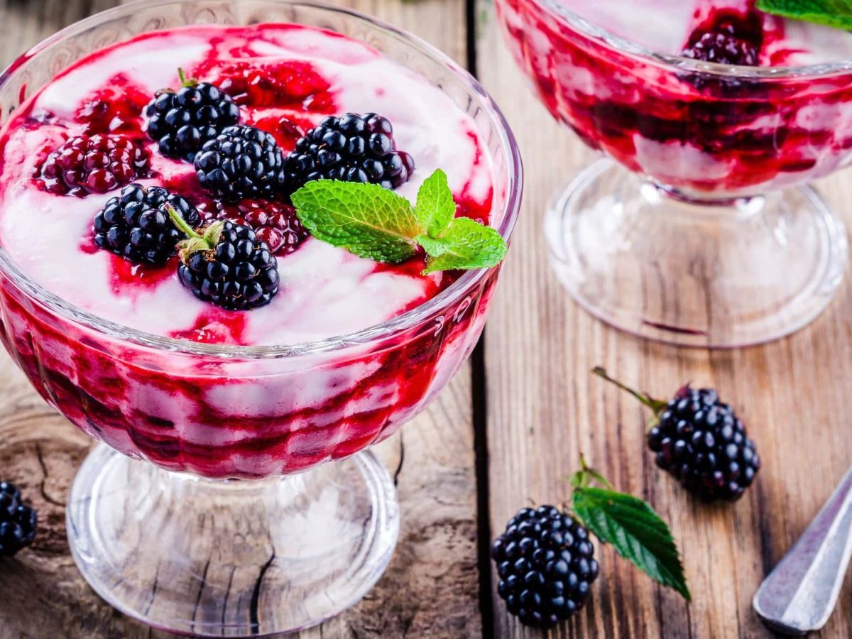 Schräge Draufsicht: Zwei Dessert-Gläser mit einer Brombeer-Sahne-Joghurt Mischung auf einem Holztisch.