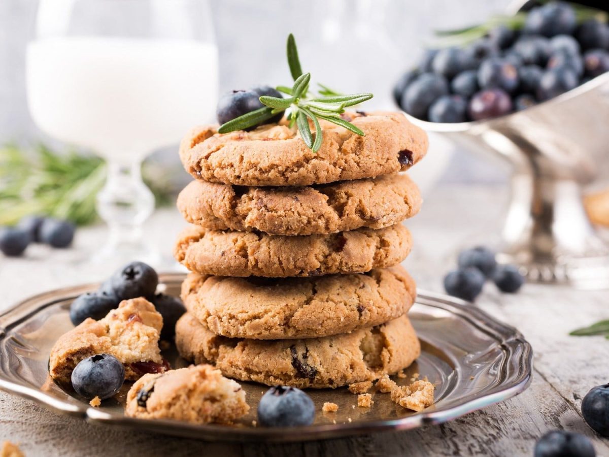 Ein Stapel Blaubeer-Cookies auf einem Teller, einige zerbrochen, daneben und dahinter Blaubeeren und ein Glas MIlch.