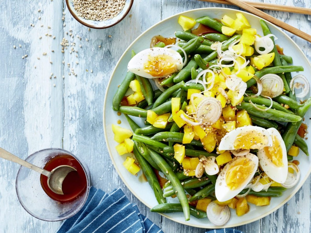 Draufsicht: Bohnensalat mit Ei und Mango auf einem Teller, daneben ein Glas mit Sojasauce unr Sesamkörner auf blauem Holzuntergrund.