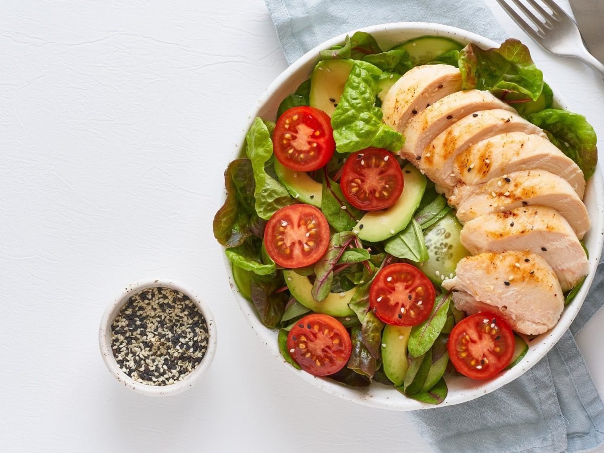 Draufsicht: Auf hellblauem Untergrund steht eine Grüne Bowl mit Putenfilet und Mandel-Dressing. Daneben lieben eine Besteck und ein Schälchen mit Sesamsamen.