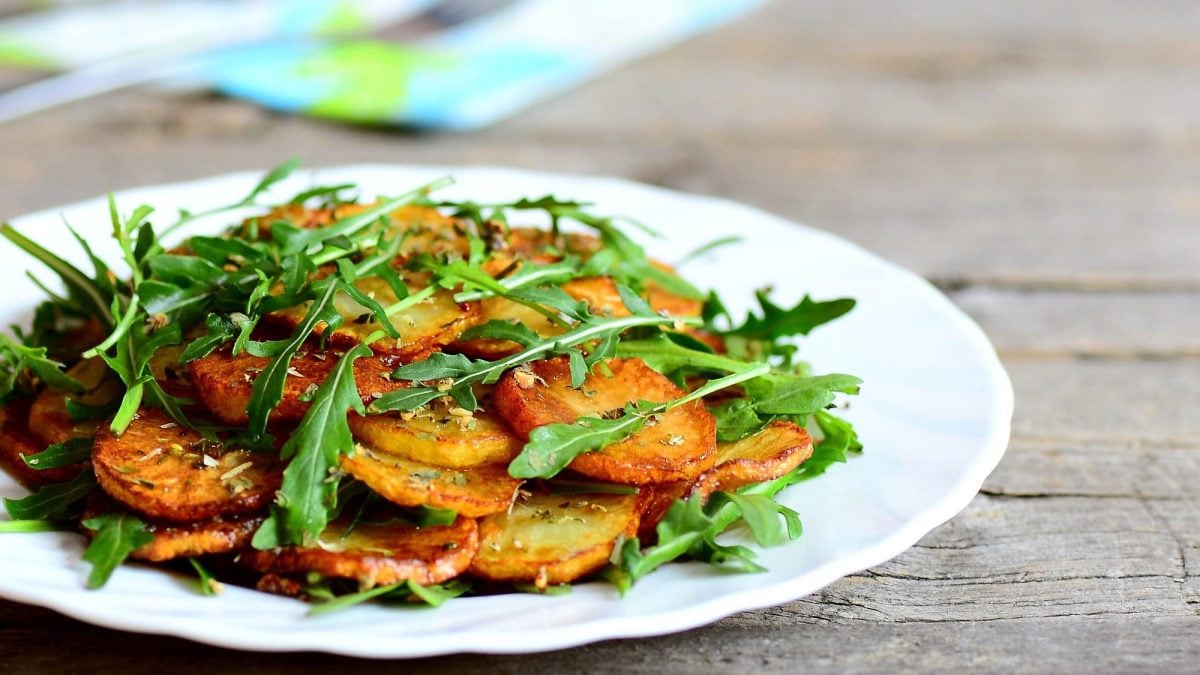 Ein Teller Bratkartoffeln mit Rucola und Knoblauch auf einem Holztisch, dahinter eine Serviette.