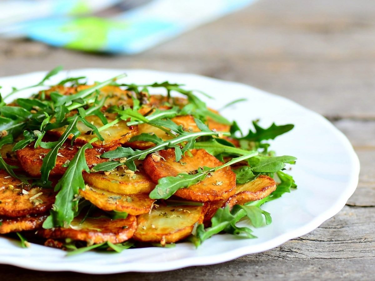 Ein Teller Bratkartoffeln mit Rucola und Knoblauch auf einem Holztisch, dahinter eine Serviette.