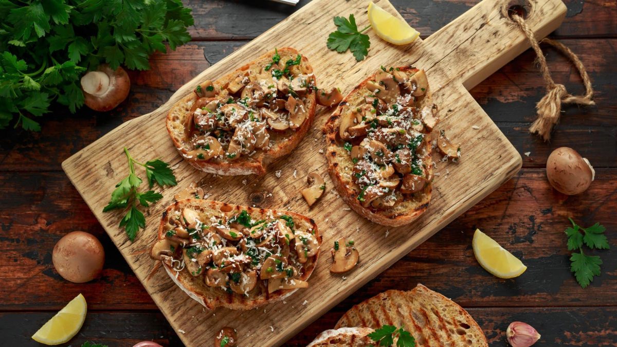 Geröstete Bratstulle mit Pilzen und Parmesan auf Holzbrett.