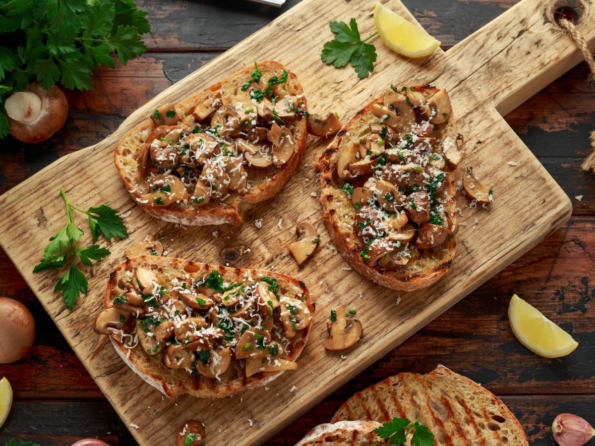 Geröstete Bratstulle mit Pilzen und Parmesan auf Holzbrett.