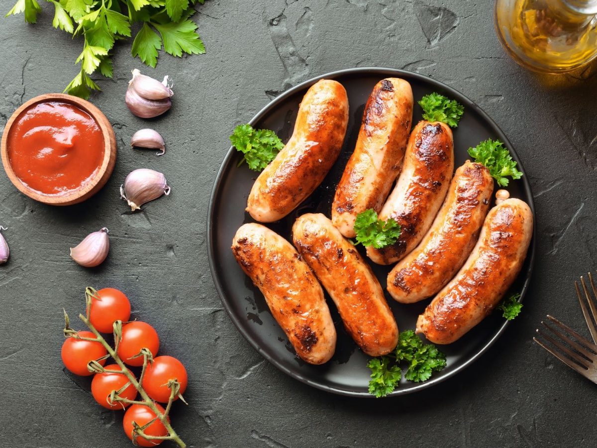 Kann man Bratwurst einfrieren? Sieben gegrillte Bratwürste mit Petersilie auf schwarzem Teller und grauem Untergrund. Im Bild Tomaten, Gewürze und eine Schale mit rotem Dip. Draufsicht.